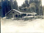 Outside a large log camp with men and empty sleds in front